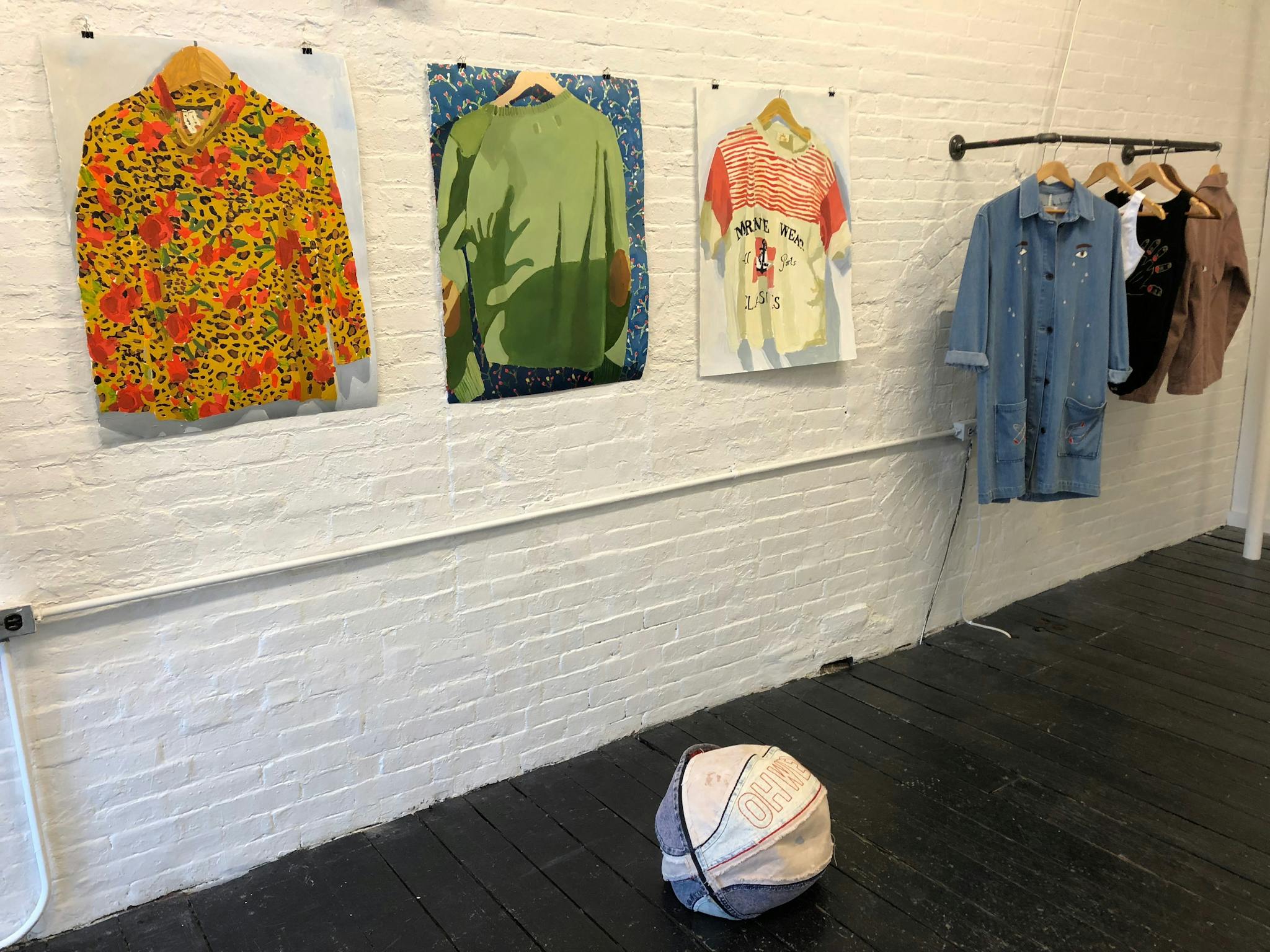 Image of one of the walls in the gallery with three paintings hung on the left and four clothing items hung on the right. Foregrounded on the floor is a basketball made out of cloth.