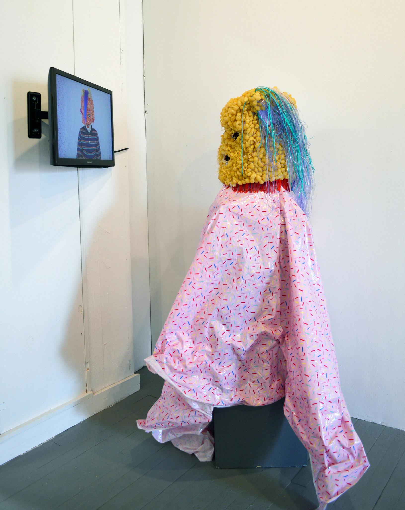 A figure wears a yellow headpiece and a pink smock, while watching a screen positioned on the wall.