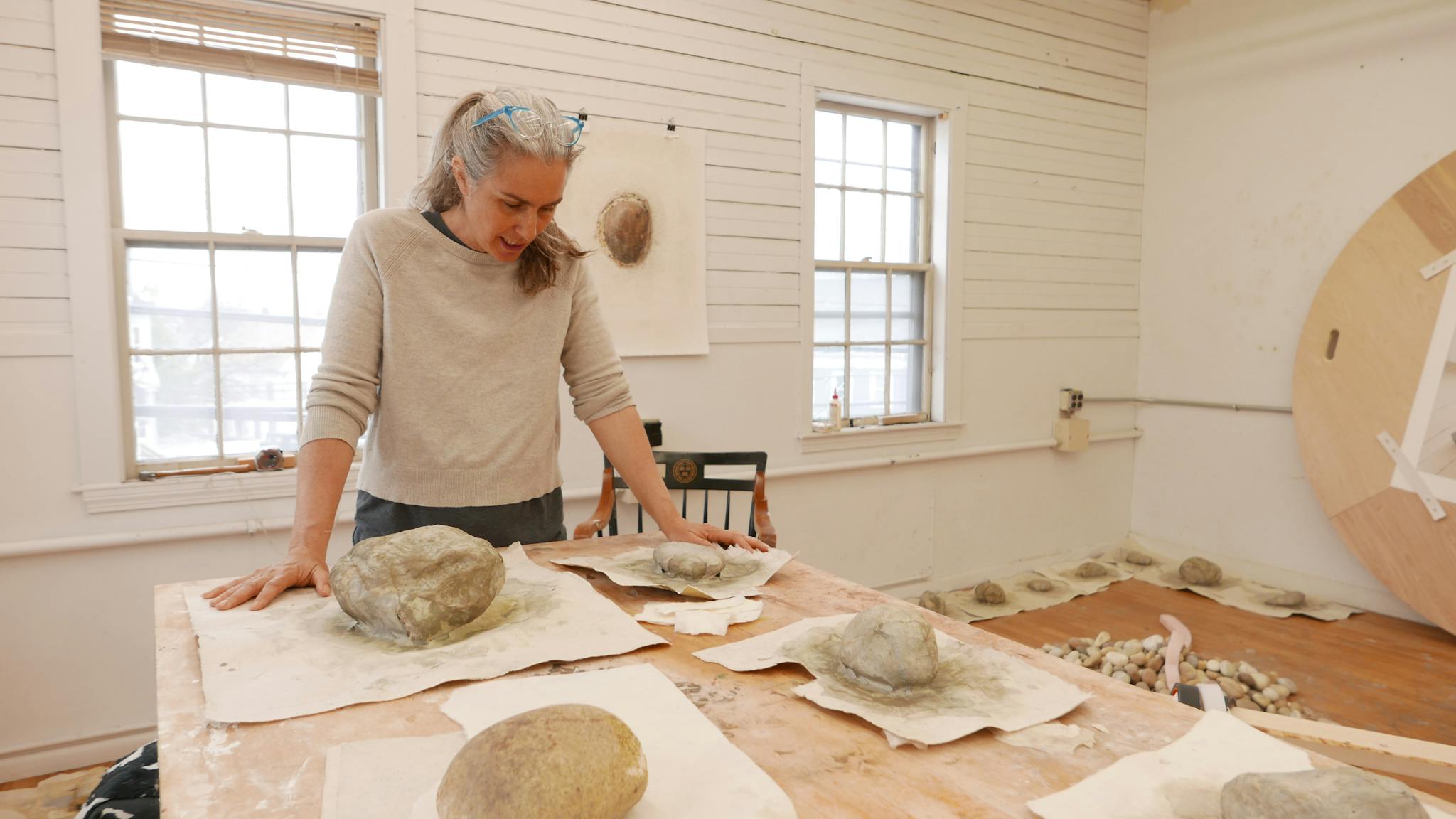 Tory Fair stands over a table in her studio, looking at sculpture works.