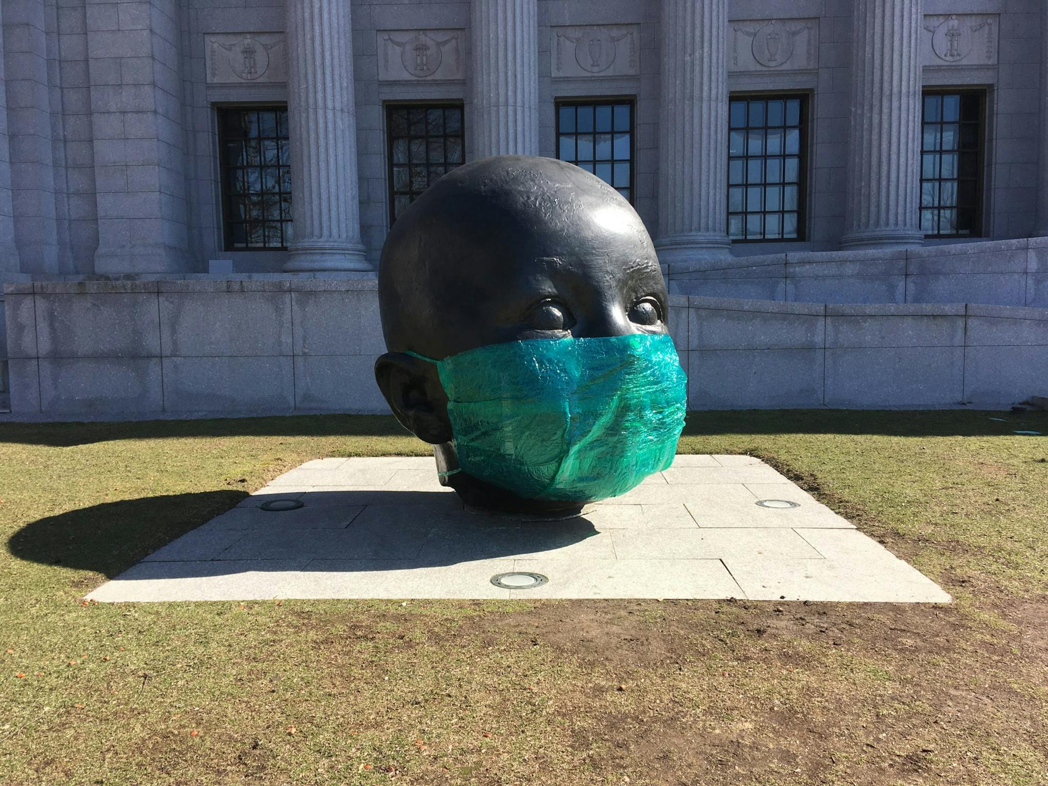 A sculpture represents an individual's head, with a green COVID-19 mask appearing on the lower half of the face.
