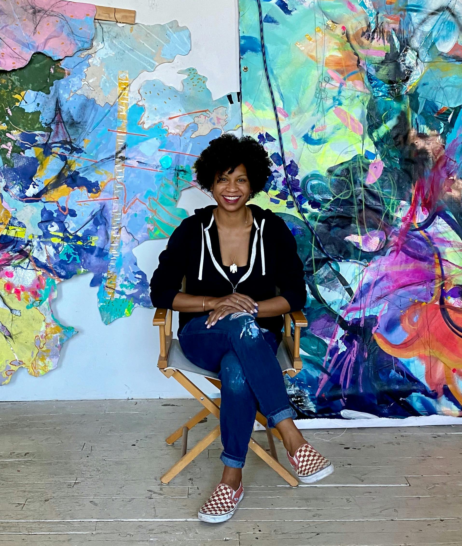Cicely Carew sits, smiling, in front of a lush, colorful work of art she has created, in her Waltham studio.