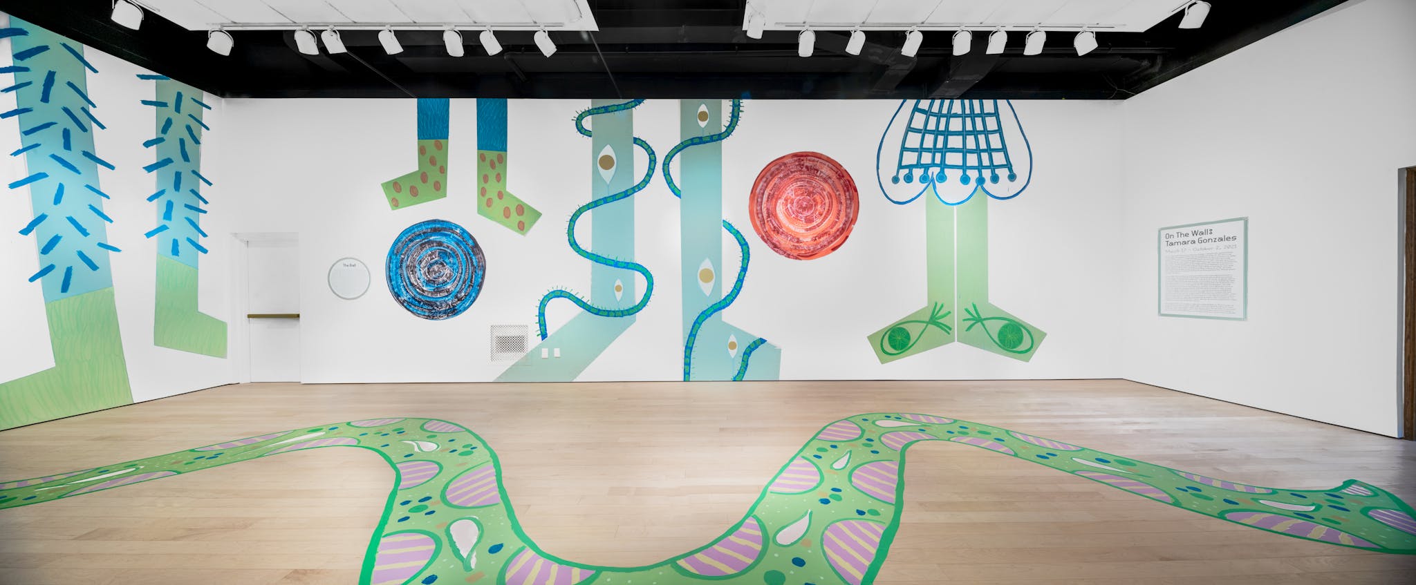 Whimsical green and blue paintings by Tamara Gonzales cover the floor and walls of the gallery space.