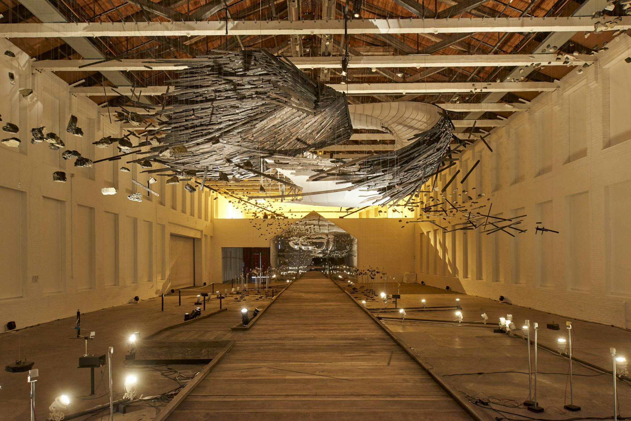 Glenn Kaino's installation displayed in a room with a wooden floor, hanging sculptural work, and a series of lights.
