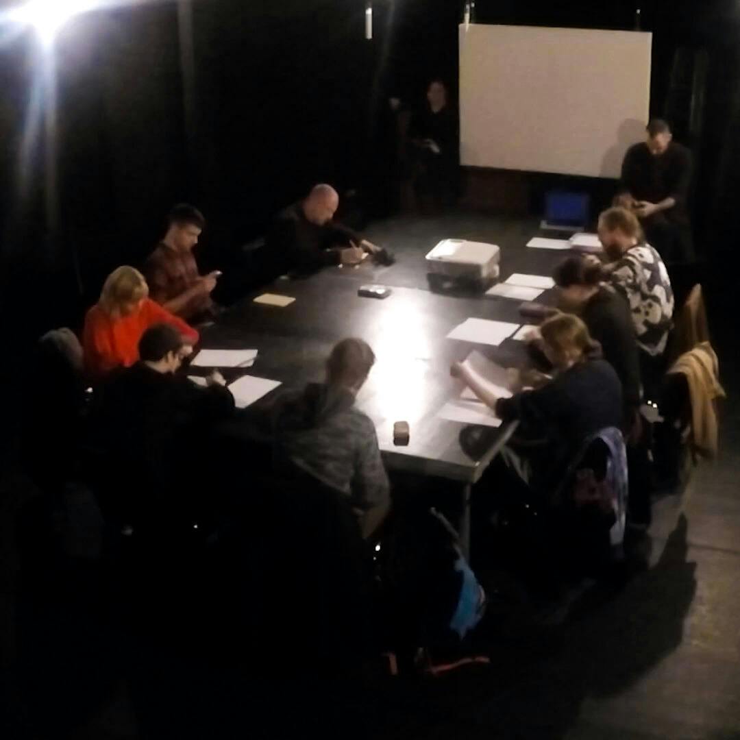 A group of people sit around a table in a darkened room, taking notes on paper.