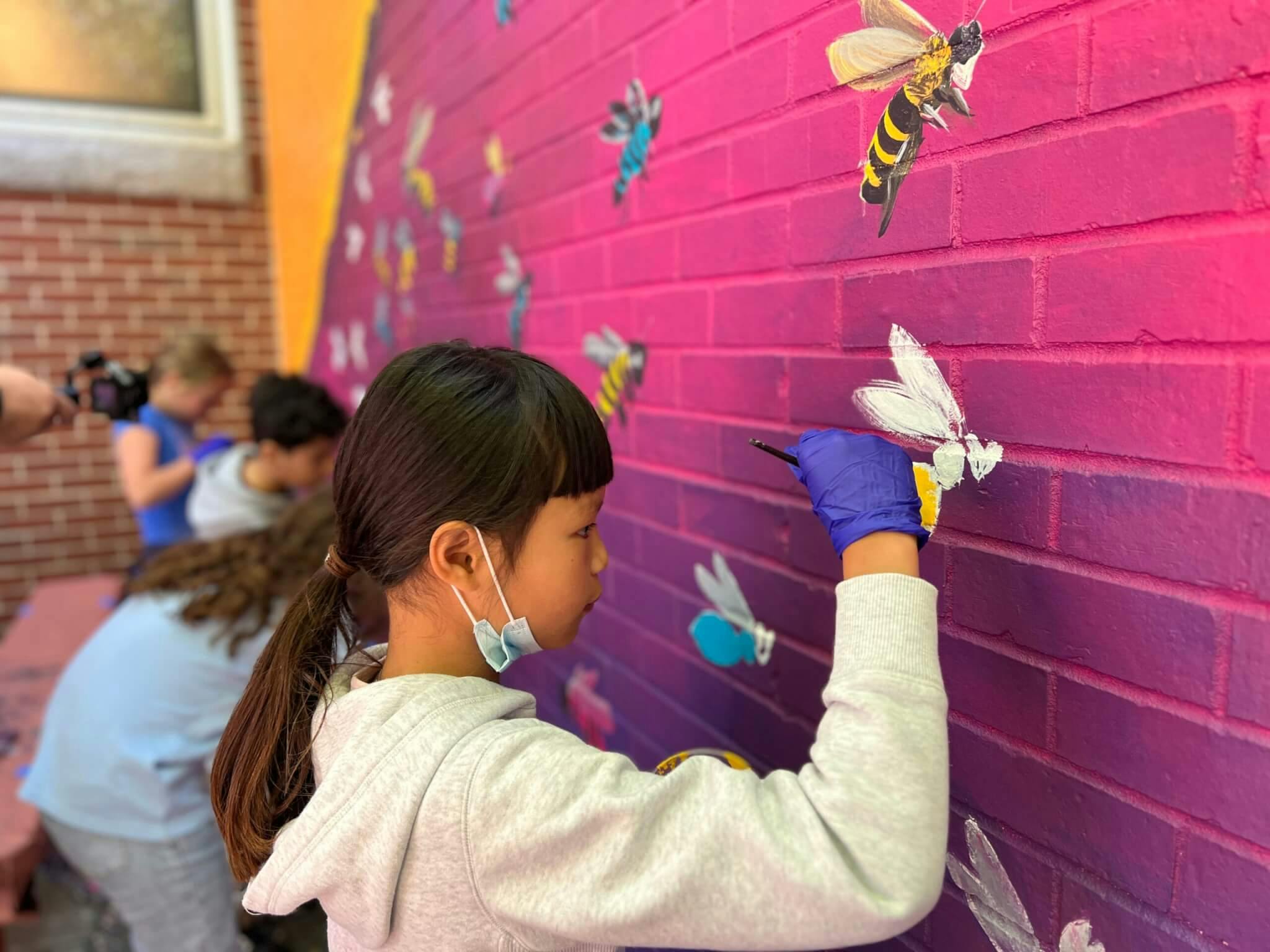 A young girl wearing a white sweatshirt paints bees on a bright pink wall.