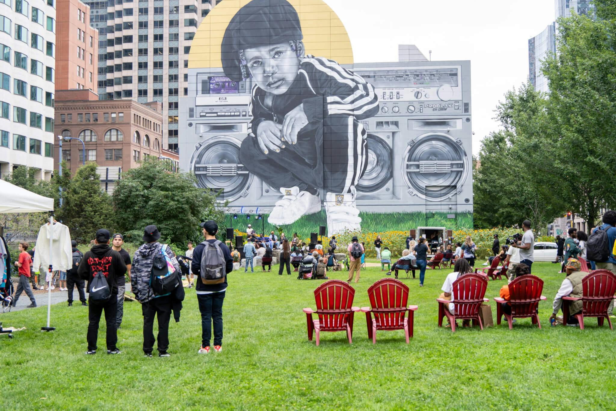 Image of “Sound In the City” event at the Rose Kennedy Greenway. People are standing and sitting on the grass in the foreground, and panelists sit in the background in front of Rob “ProBlak” Gibbs' "Breathe Life" mural
