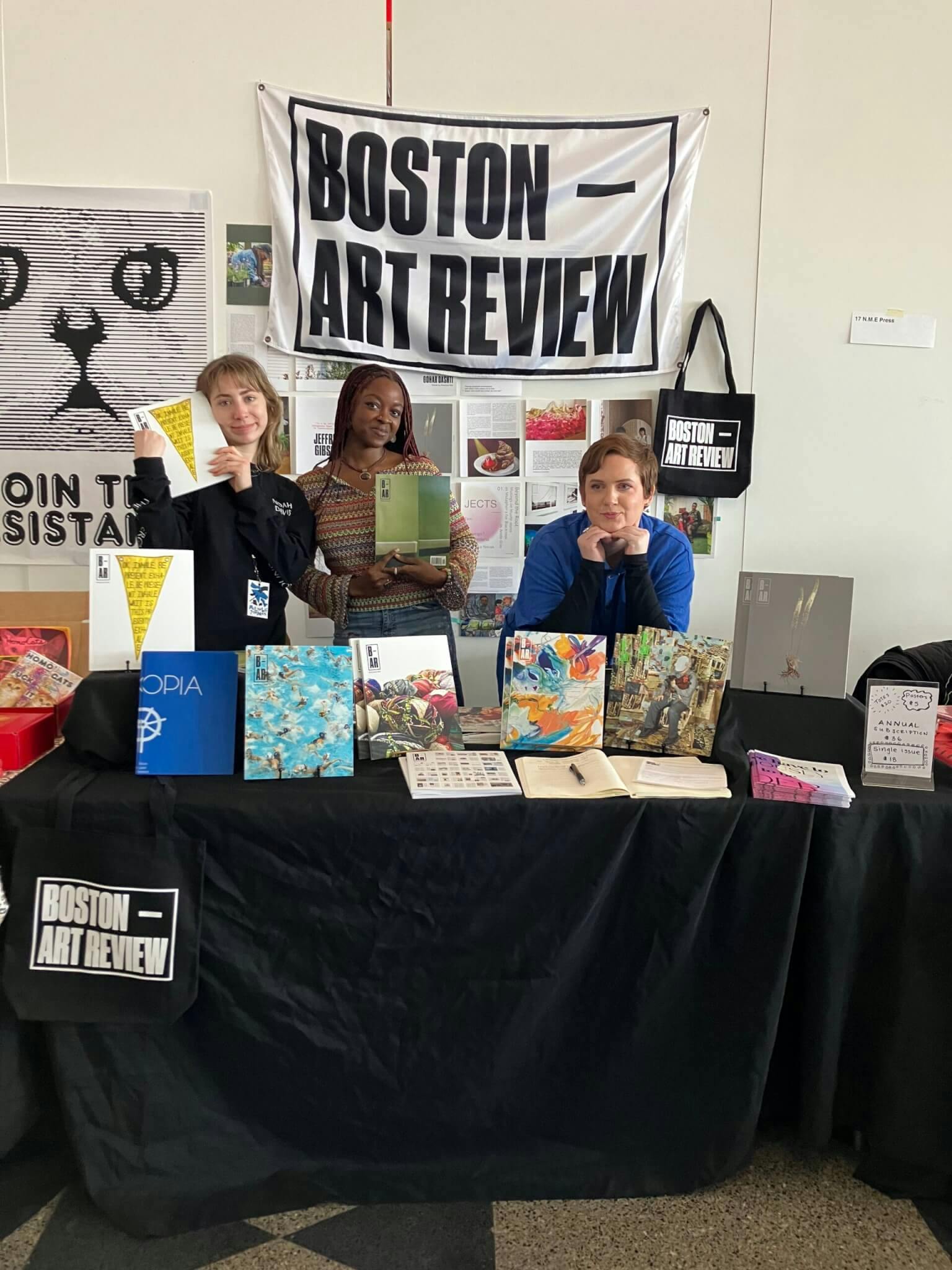 Three of the Boston Art Review team tabling at the 2023 Multiple Formats book fair. They pose behind a table that flaunts many previous issues, and beneath a Boston Art Review flag.