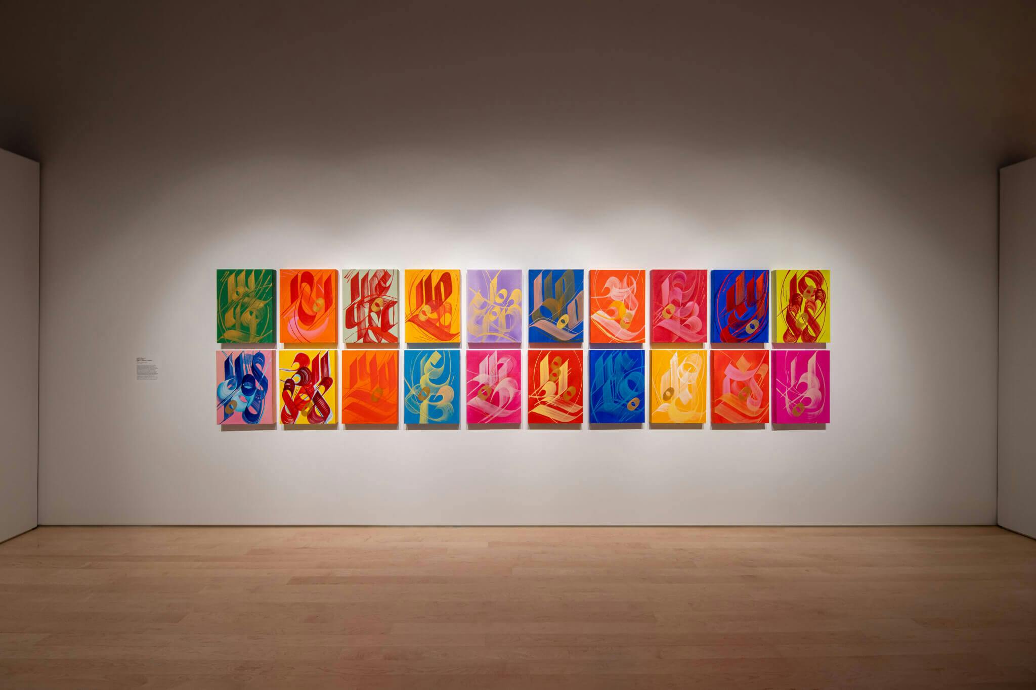 Two rows of 10 brightly colored works are displayed on a white wall with minimal lighting.