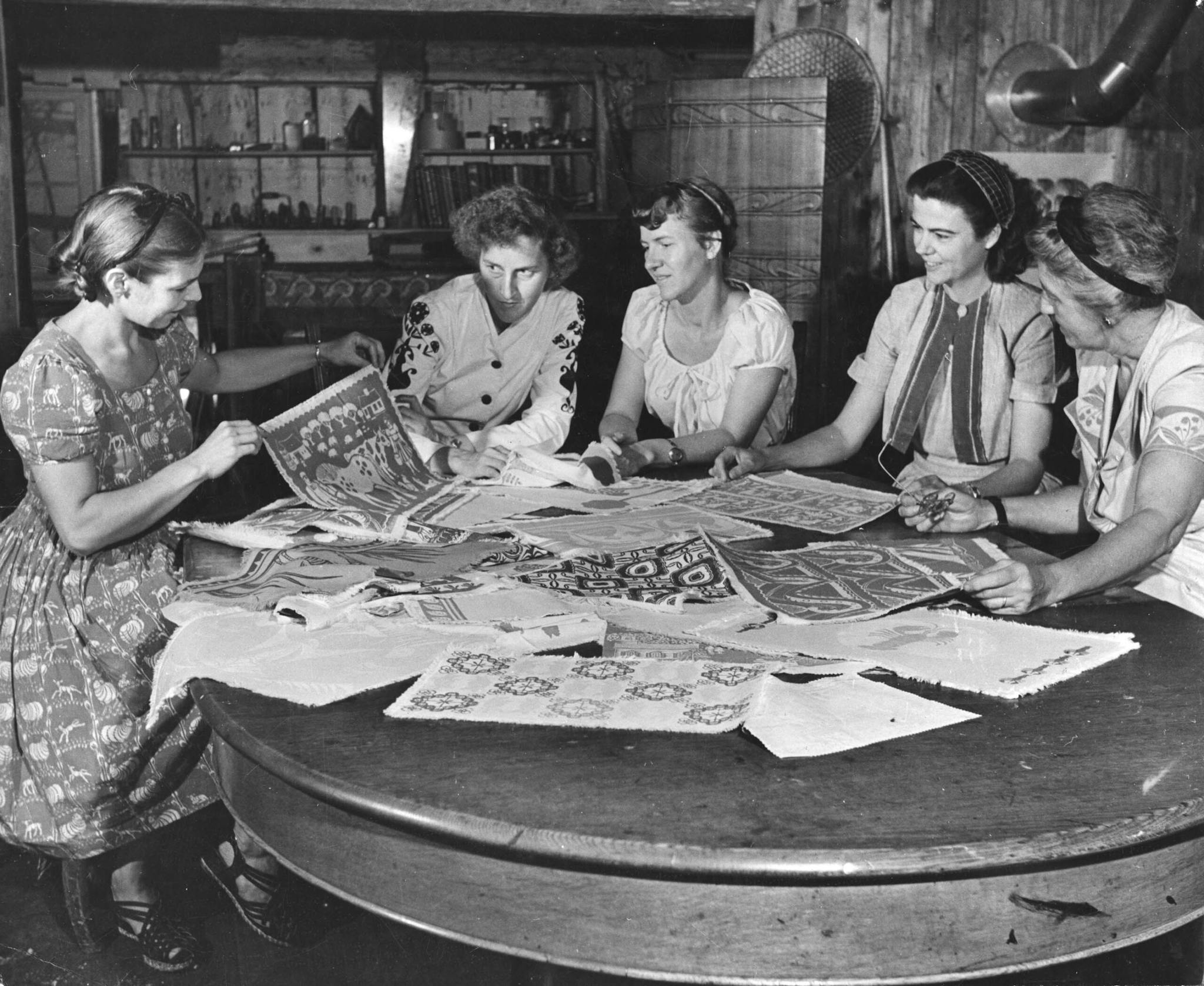 A black and white photo of a group of women seated at a table where designs are laid out.
