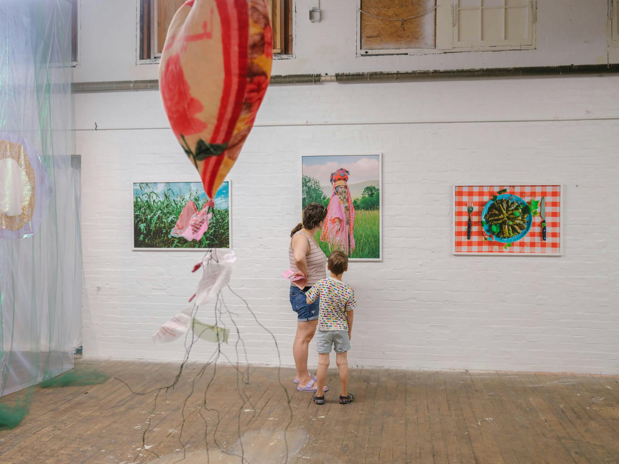 An installation view of the exhibition. A mother and her child look at the center of three works displayed on a white wall. In the foreground is a spidery red and orange sculpture.