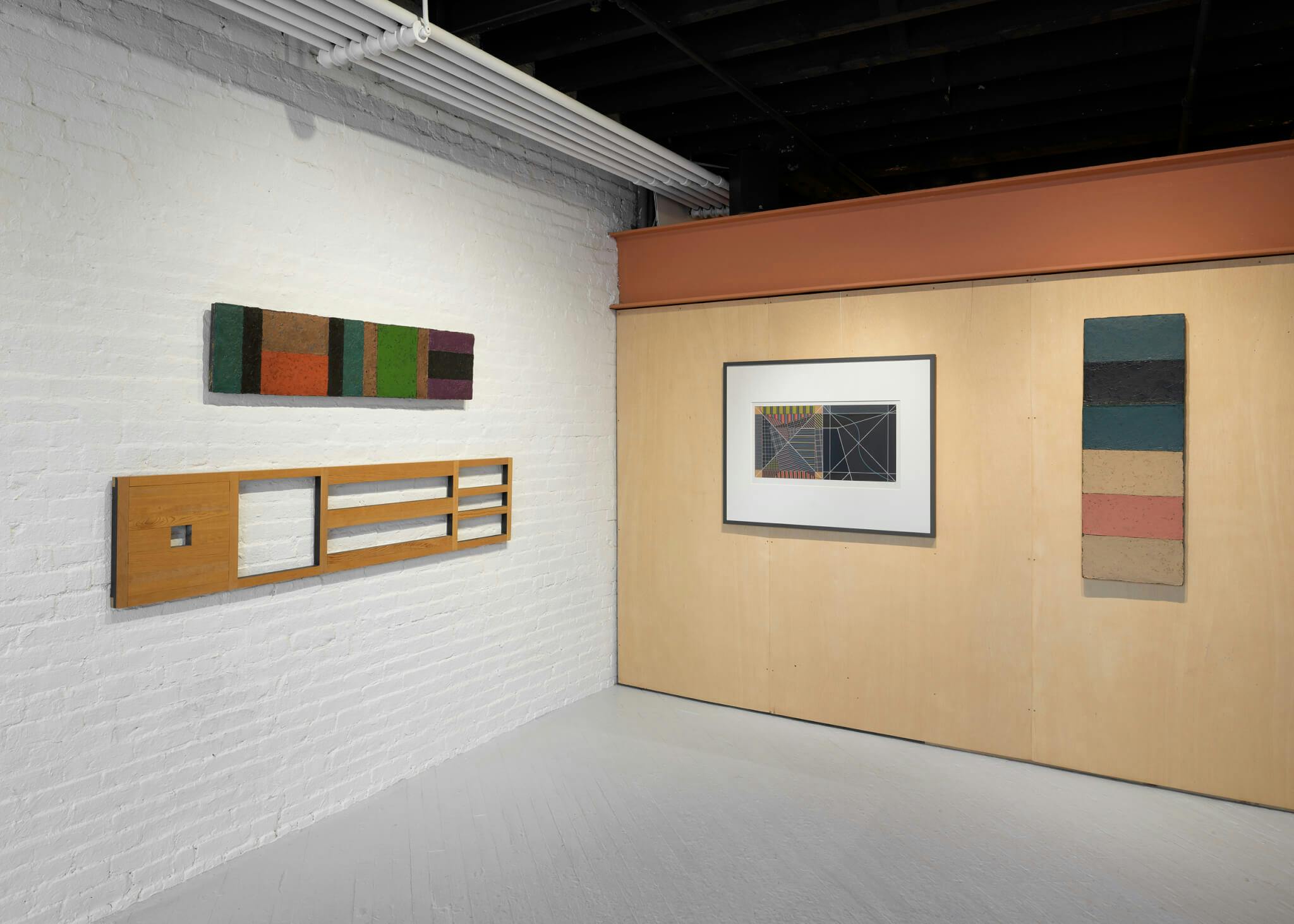 An installation view of the Rob Moore Retrospective. The image is a view of a corner -- on the white wall to the left are to rectangular works, and on the light-brown wall to the right is a framed piece next to another rectangular work.
