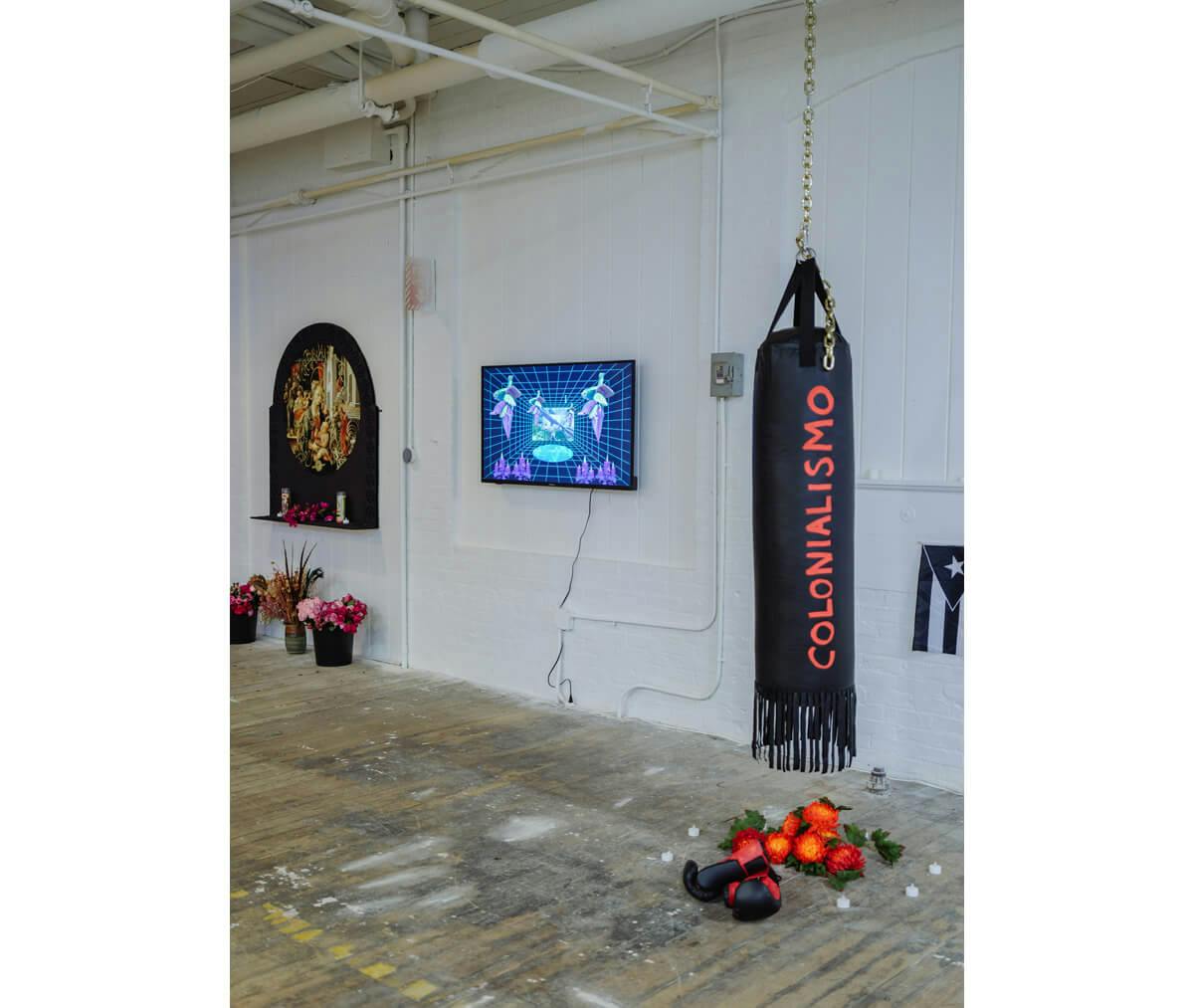 An installation view of the exhibition. To the right hangs a black punching bag with the word "colonialism" written on it, in the center hangs a TV displaying something, and to the left is an arch-like work with flowers beneath.