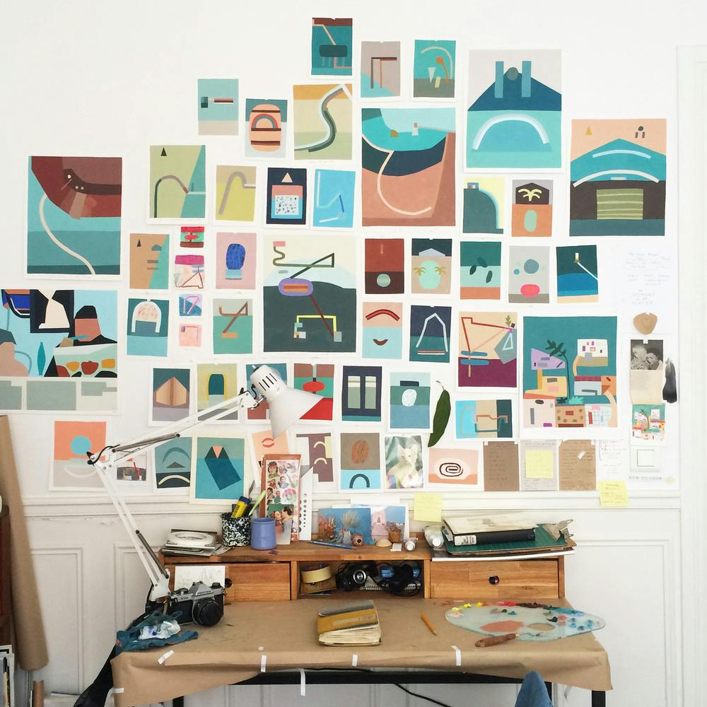 Image from Kristin Texeira's studio. A desk with painting materials and other desk objects is foregrounds a large wall full of the artist's colorful paintings.