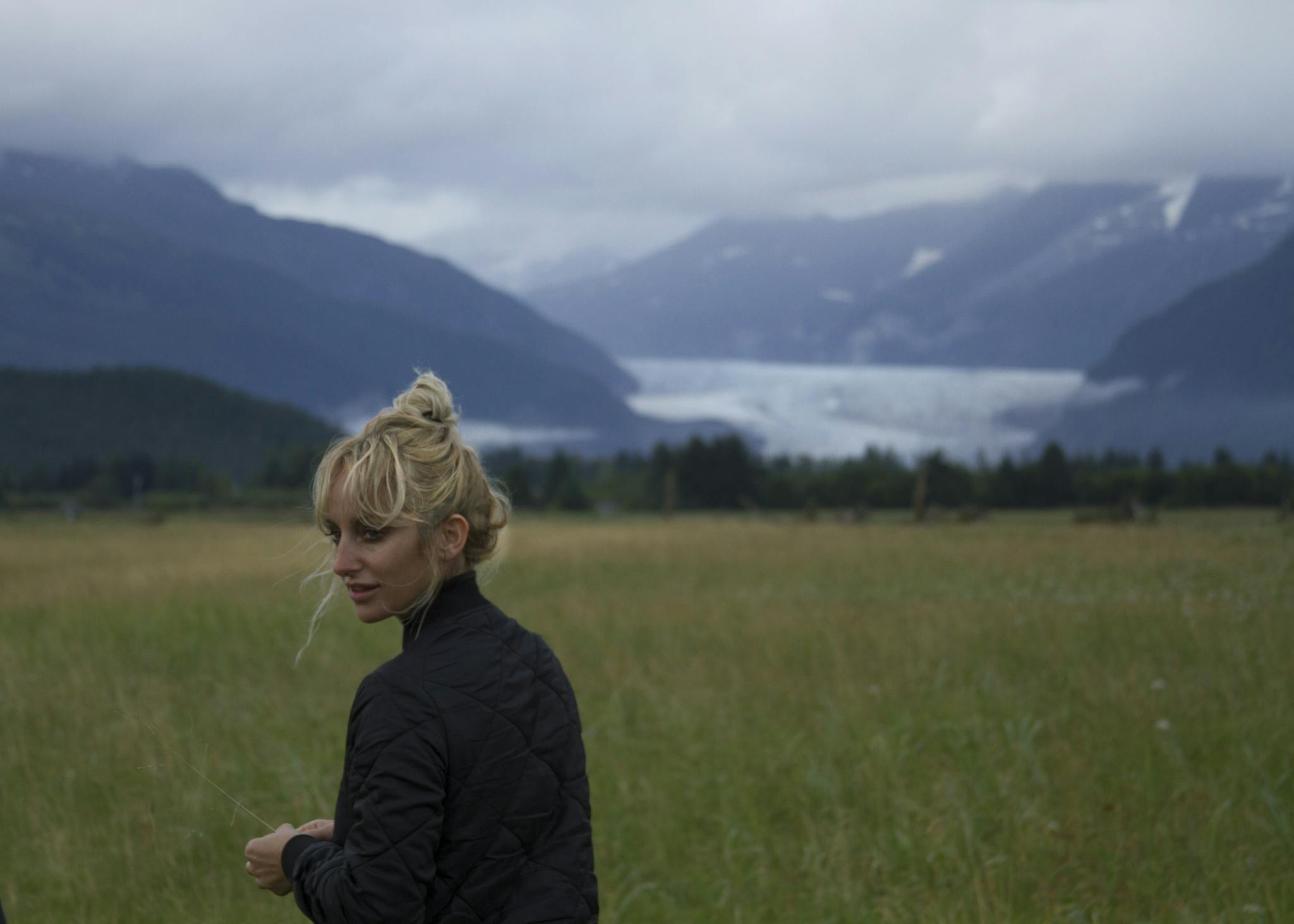 A portrait of Kylie Manning, standing in a field, with a view of mountains, behind.
