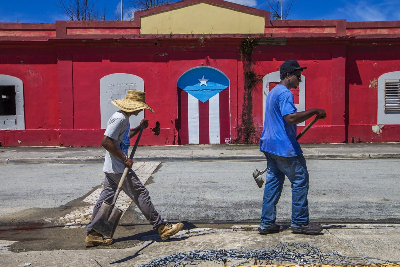 Beach workers carrying tools walk outside a bright red post office building.