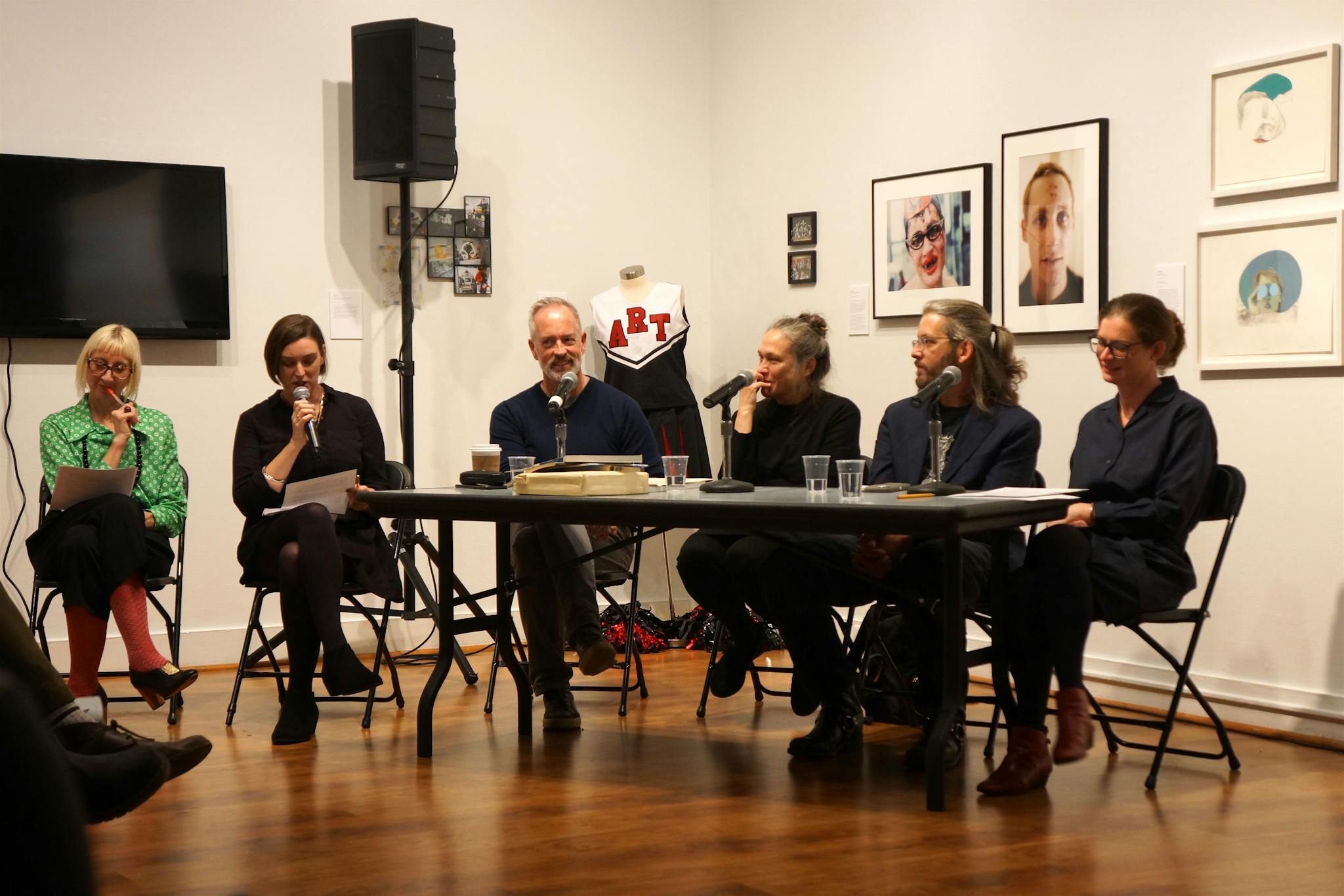 Panelists sit around a table, speaking into microphones, while artworks appear on the walls behind.
