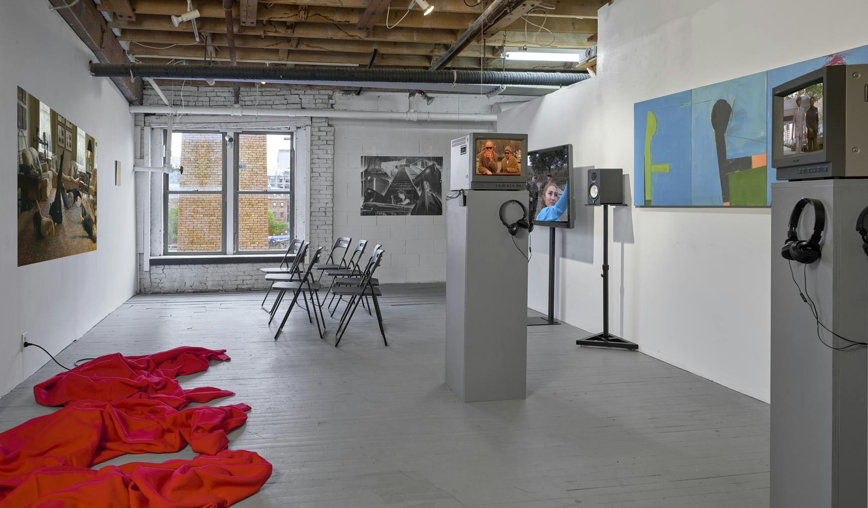 An installation features a red cloth on the floor of the space, television screens, and art hanging from the walls.