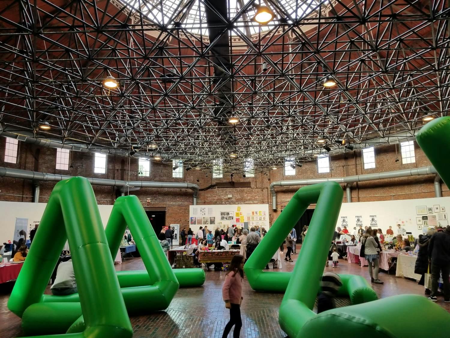 Visitors explore the Boston Art Book Fair at the Cyclorama, where large, green, inflatable structures are on display.