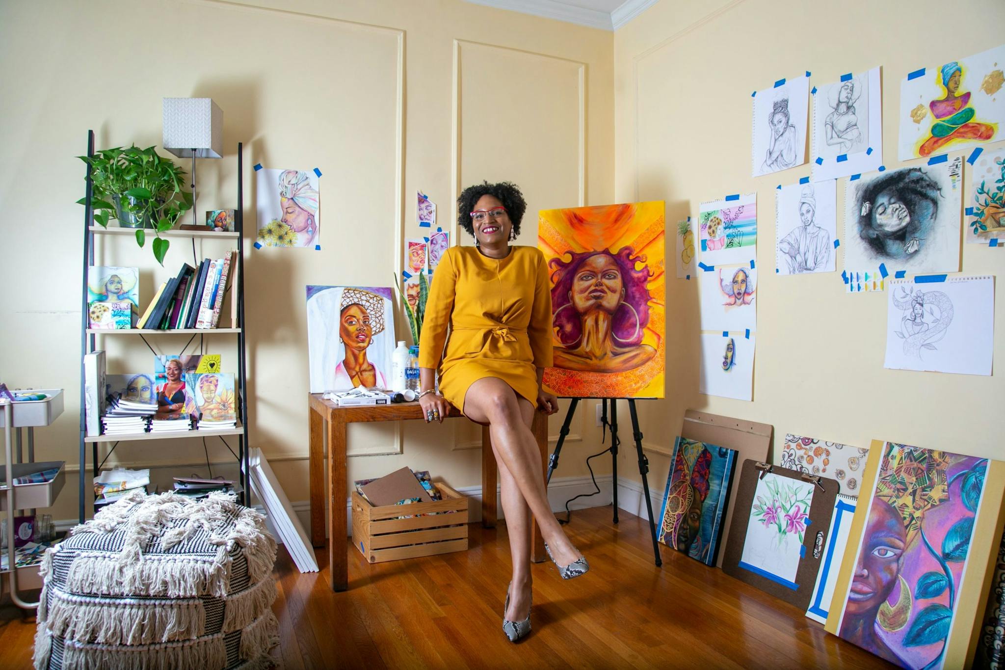 Ayana Mack is at her home studio, seated beside two paintings of women's faces, while a collection of artwork appears on the wall to the right.