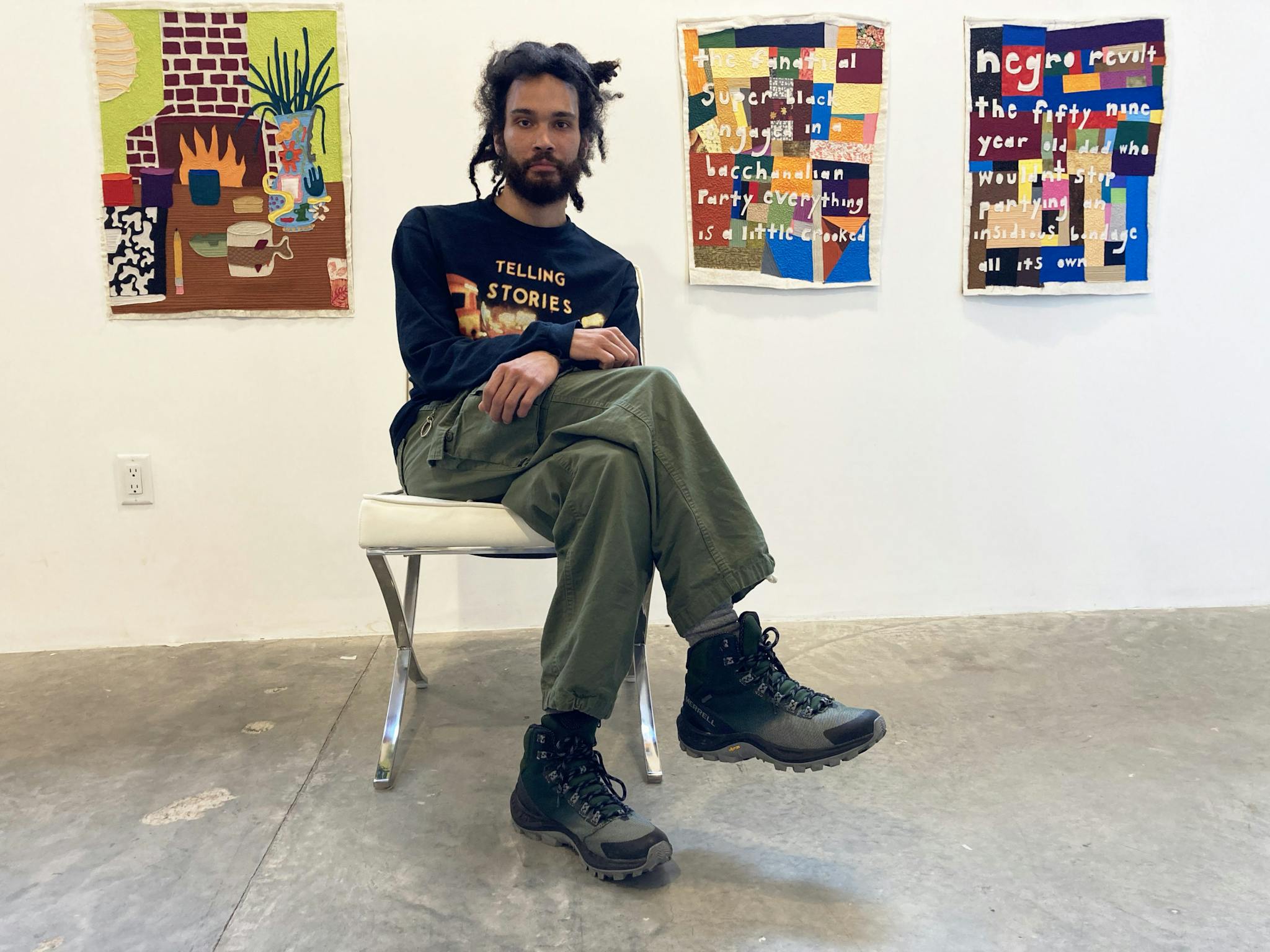 Michael C. Thorpe sits cross-legged in front of brightly colored quilted designs, hanging from the wall.