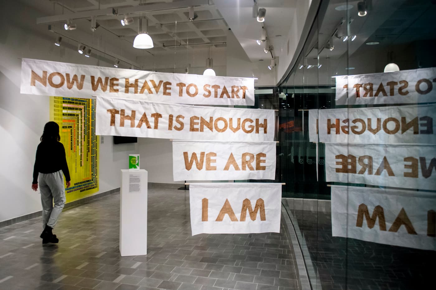 A series of signs hang from the ceiling displaying messages, while a woman walks pass them on the left hand side of the photo.