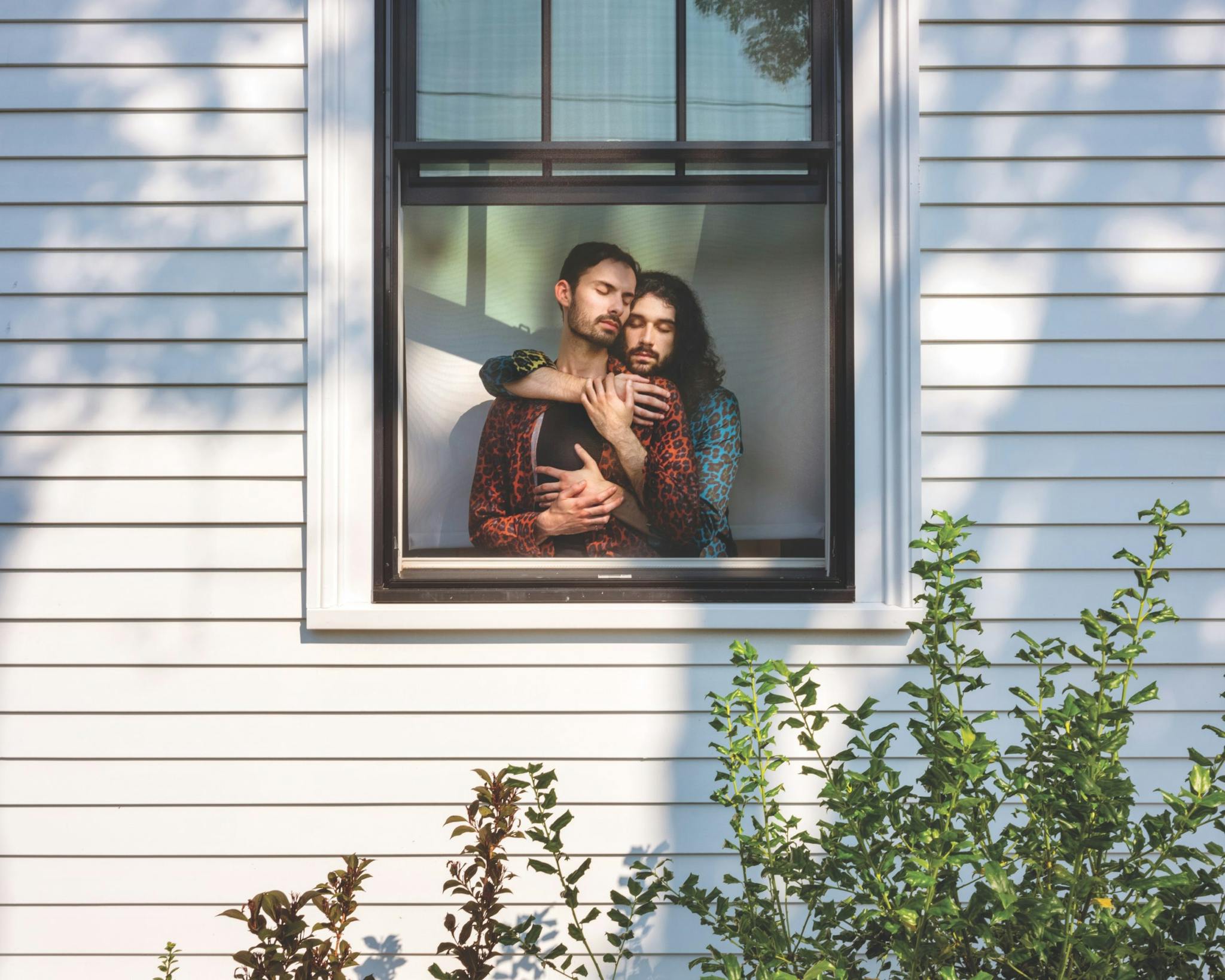 A couple named Cam and Jared pictured in the window of a house, a green plant appearing below.