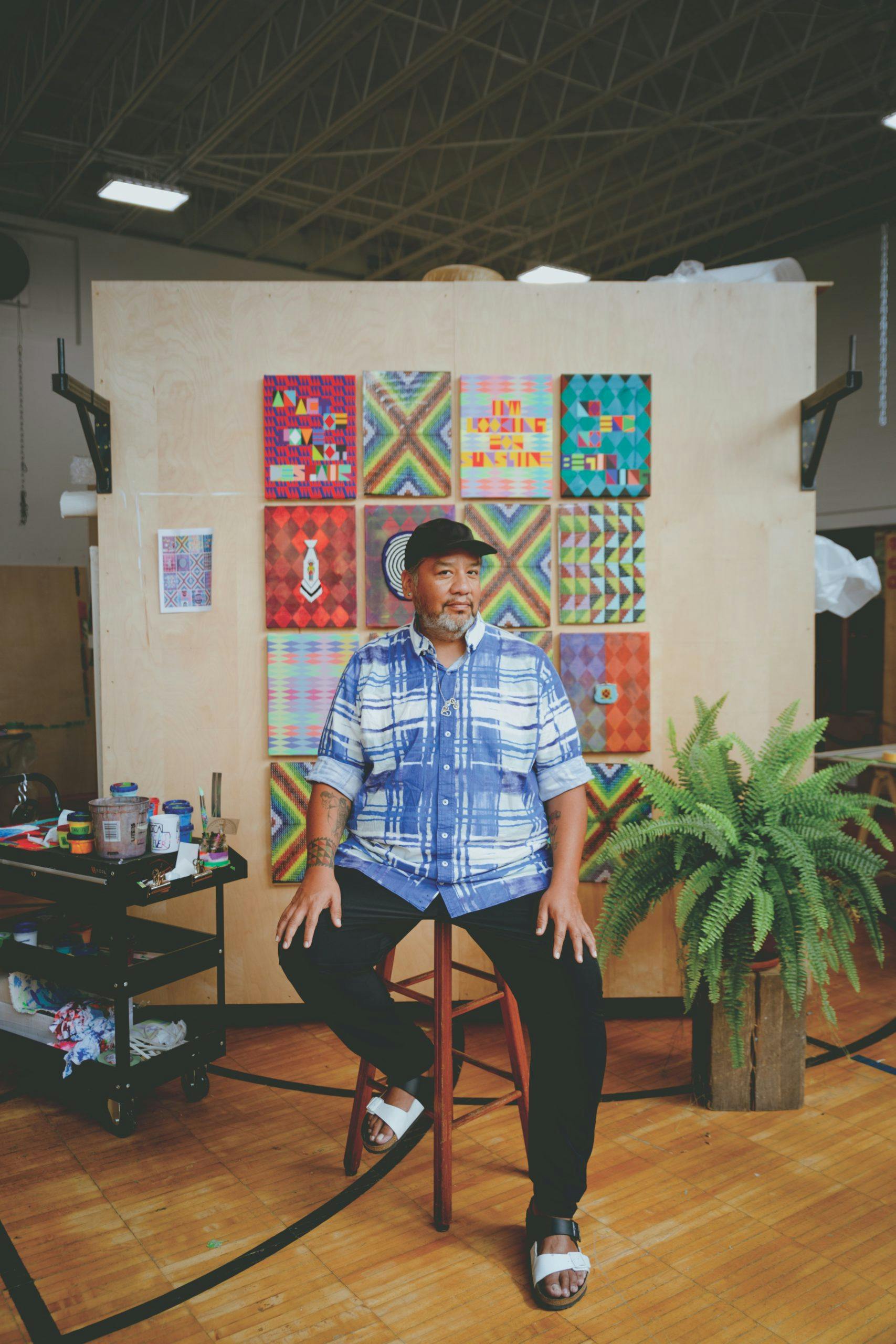 Jeffrey Gibson sits in his studio in front of colorful paintings beside a decorative plant.