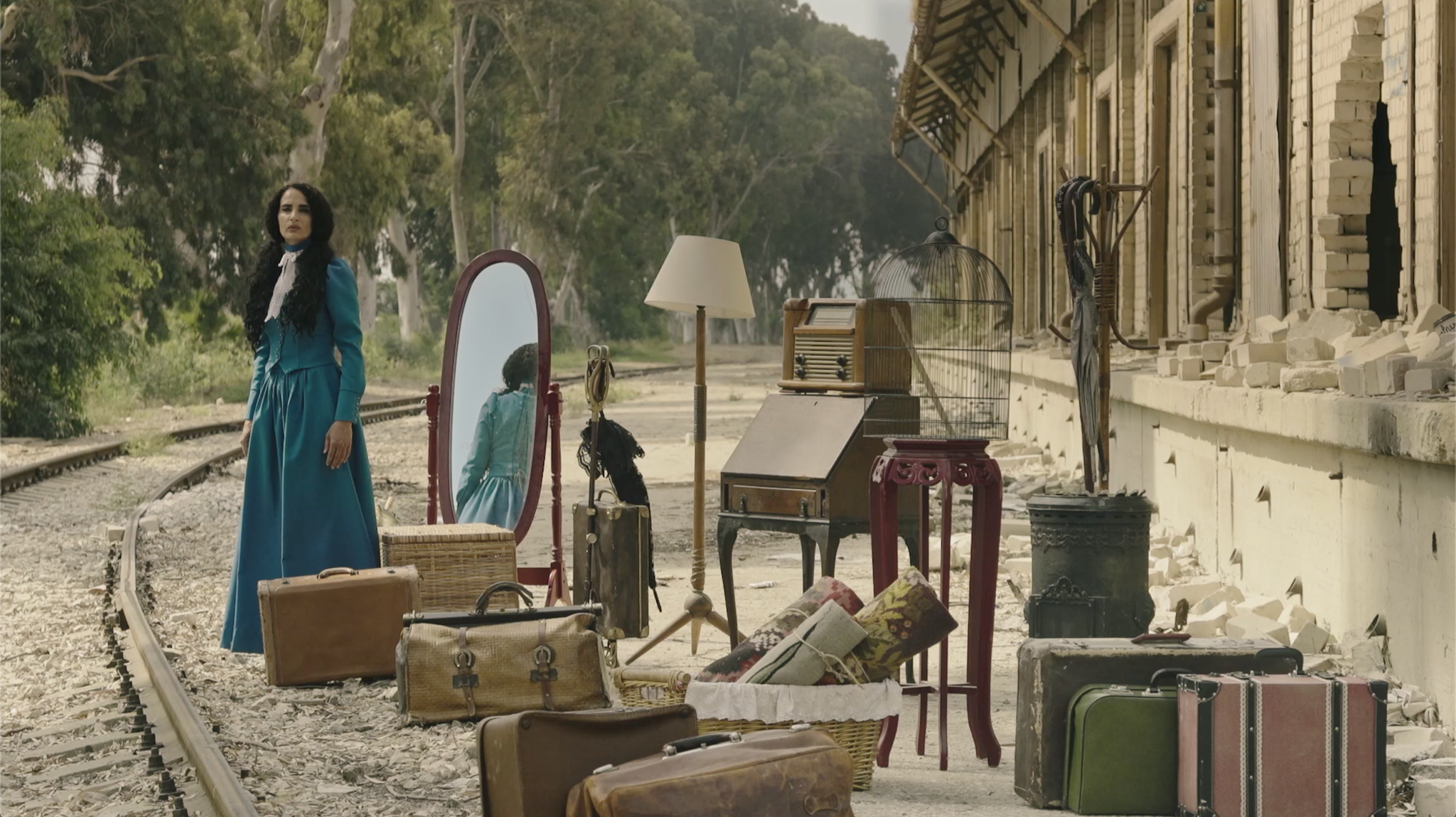 A woman in a blue dress stands beside an assemblage of furniture in the outdoors.