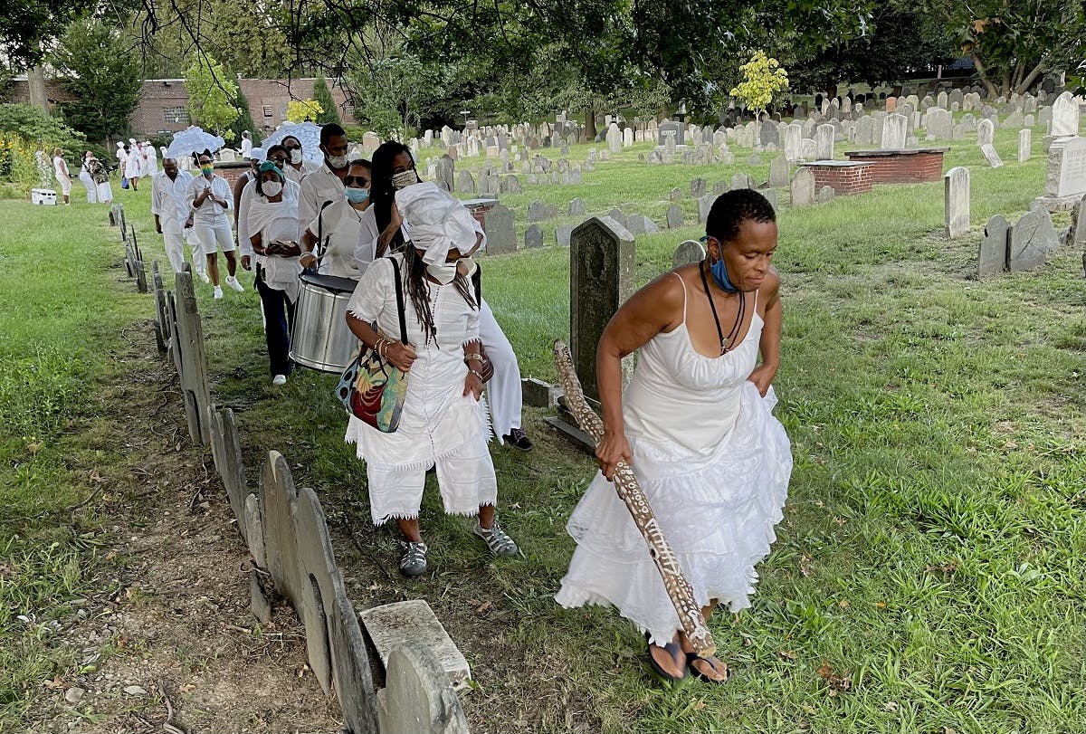 A procession of figures wearing white journey across a graveyard.