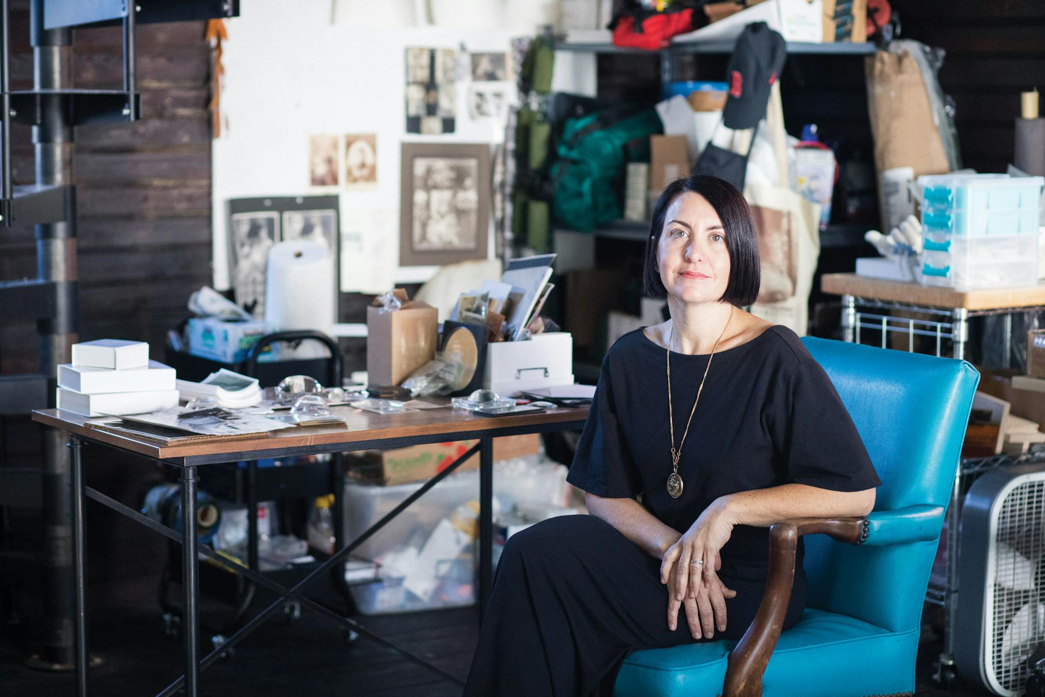 Toni Pepe inside her Malden studio, seated in a turquoise chair in front of a cluttered table.