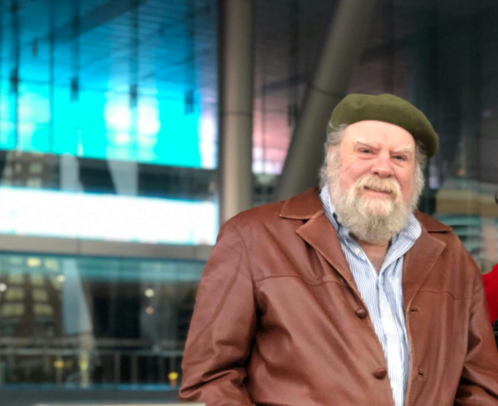 George Fifield stands in a brown jacket and green hat in front of the Boston Convention & Exhibition Center, lit by blue neon light.