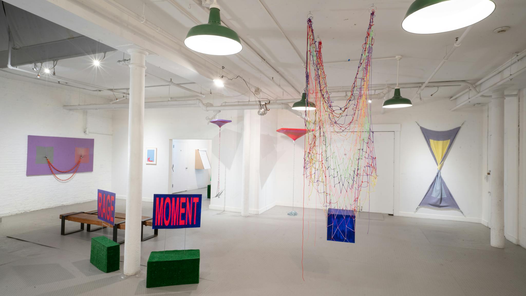 A rainbow net hangs from the ceiling, while signs display the words "rage" and "moment" in an exhibition space.