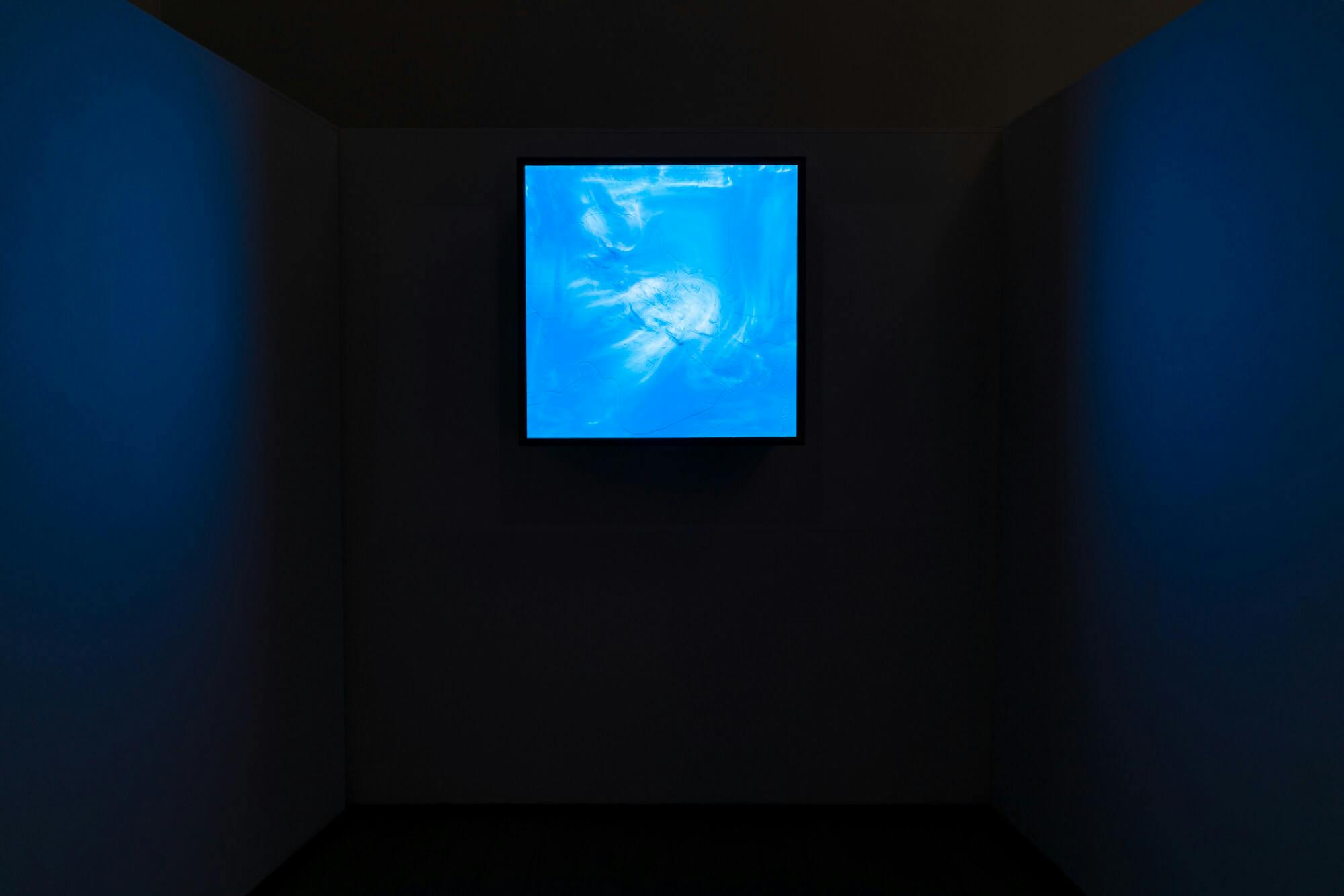 Installation image of "You Are Here." Small, bright blue square in a dark room on a pitch black wall. The blue light reflects on the side walls creating a navy hue.