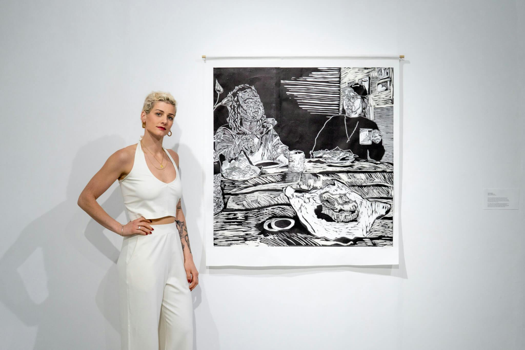 Artist Brooke Stewart, wearing white, stands beside black and white artwork positioned on the wall.
