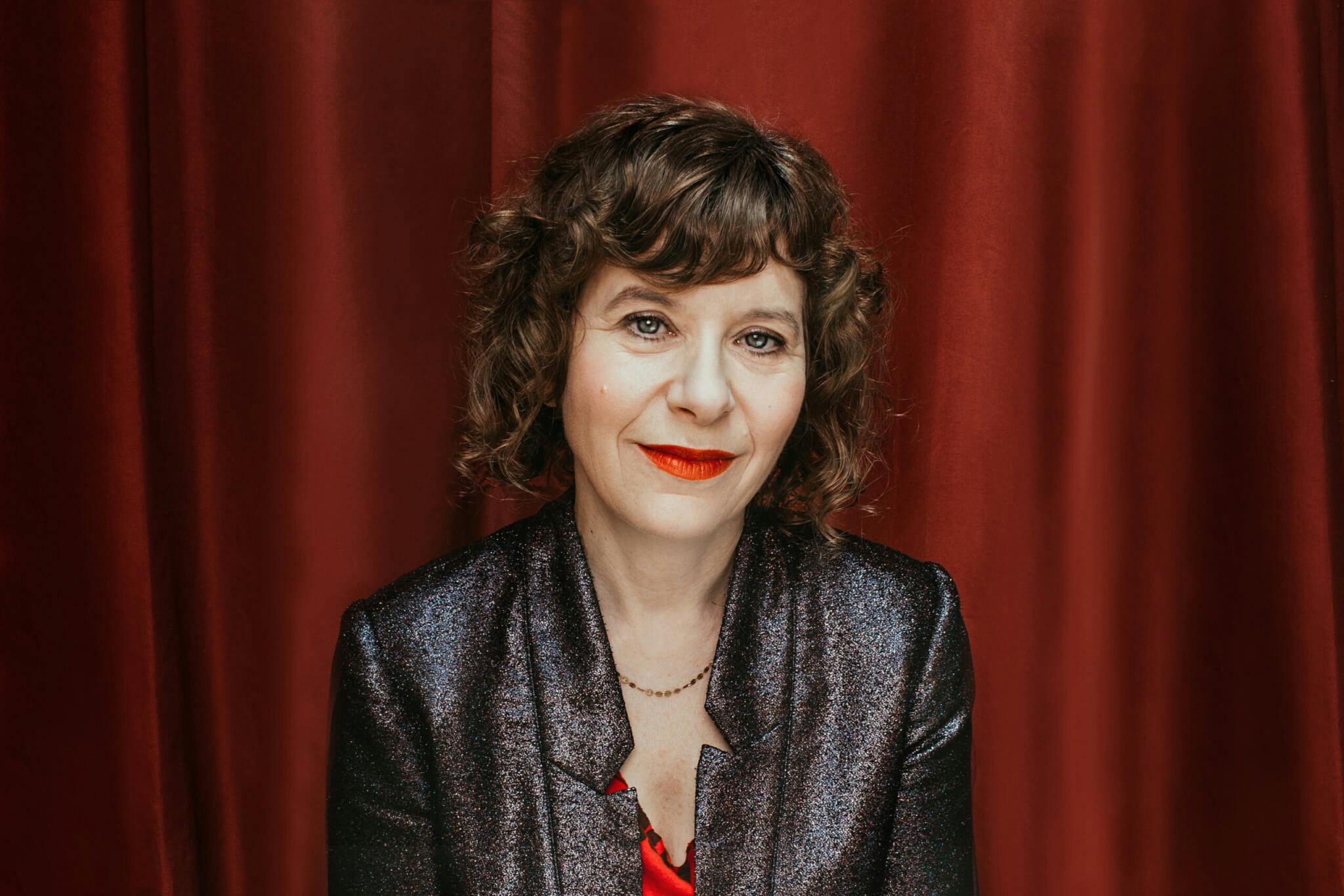 A portrait of Elisabeth Subrin, set against a red curtain.