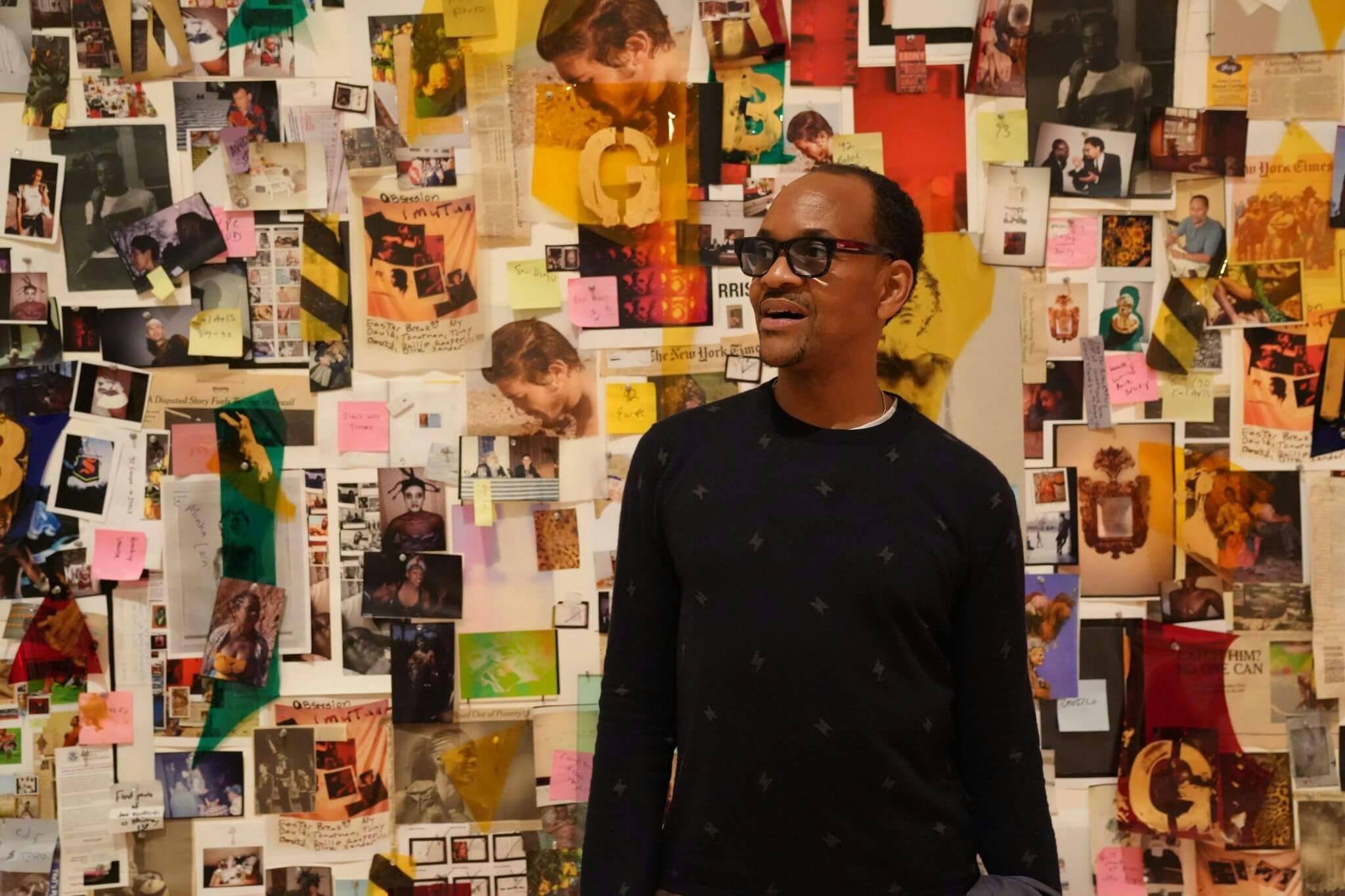 Lyle Ashton Harris wears a black shirt and glasses, standing in front of a wall of colorful images.