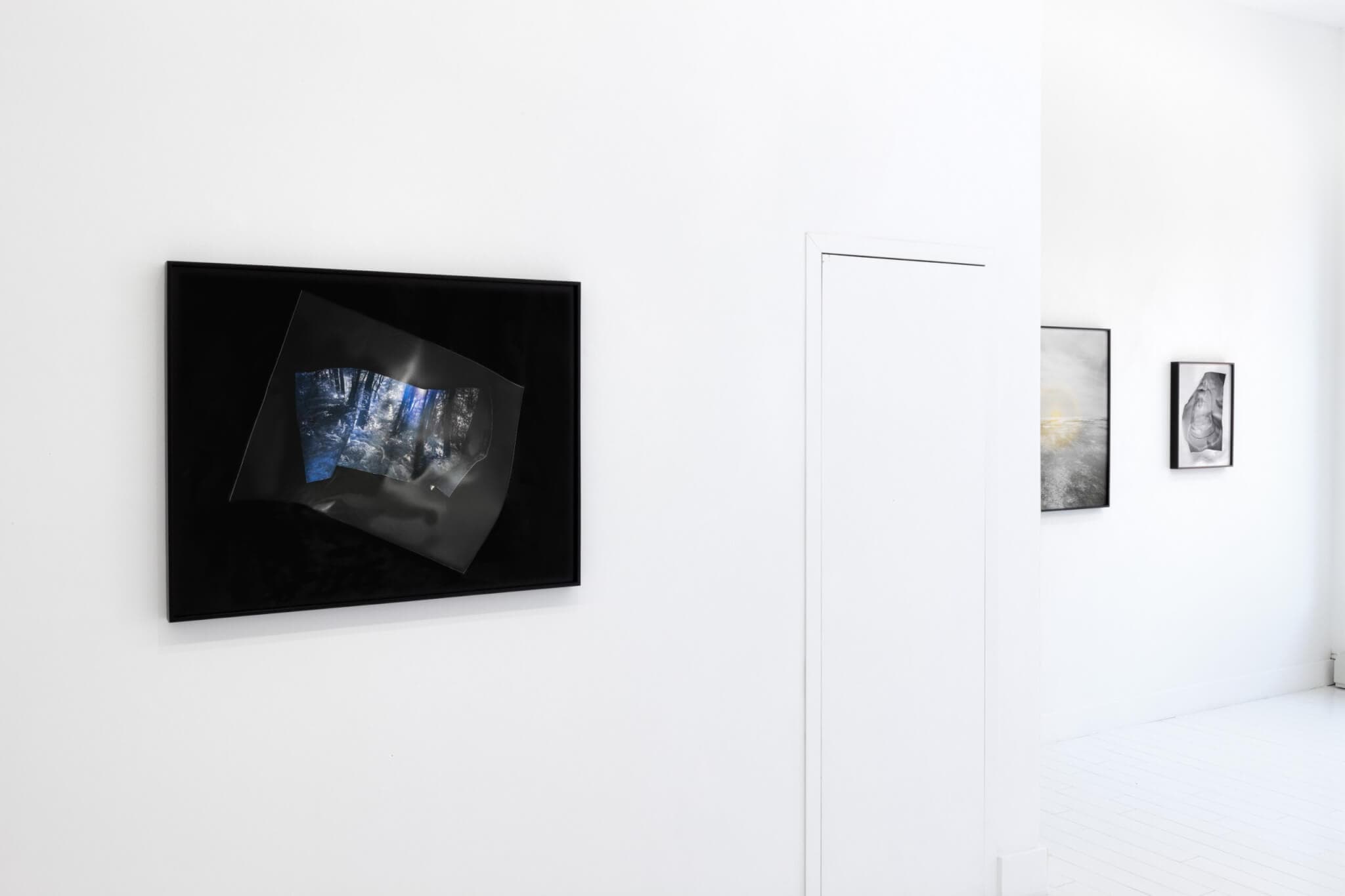 A dark photograph, hanging against a white wall, by Kate Greene features a shimmering display of light.