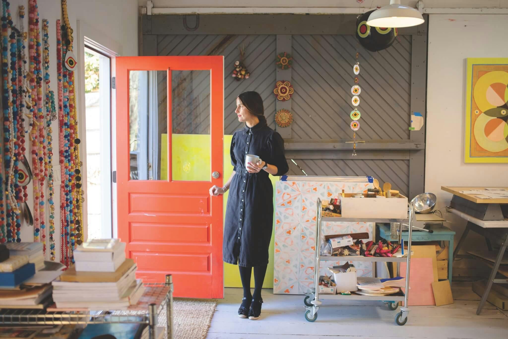Artist Crystalle Lacouture stands beside a brightly colored door in her Wellesley studio, holding a mug.