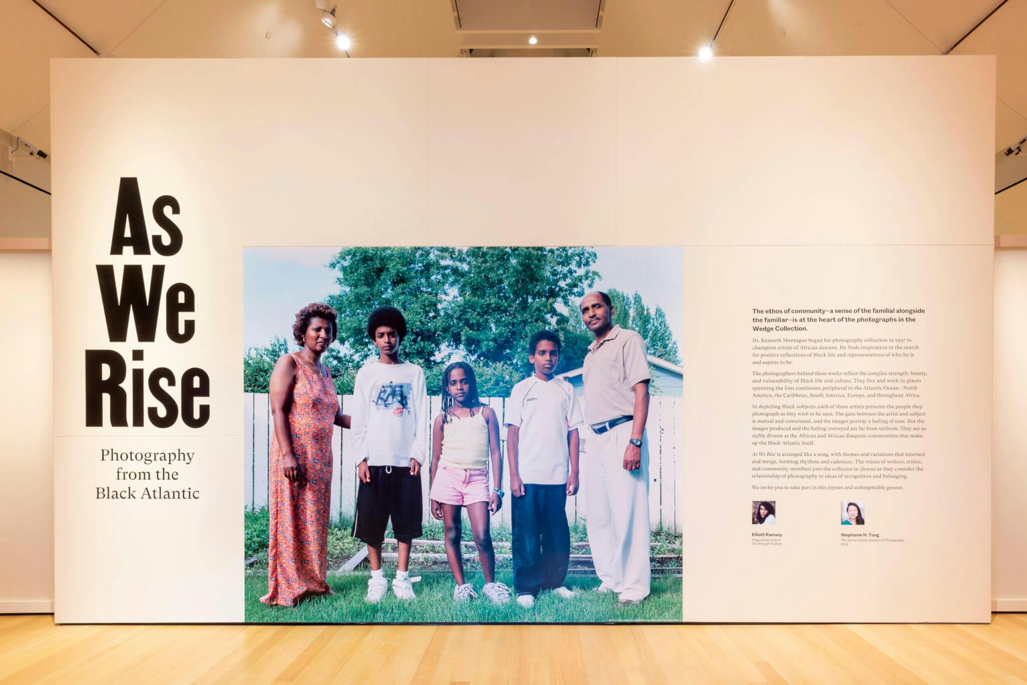 A white wall with "As We Rise" in bold font to the left, an enlarged photograph of five members of a Black family in the middle, and a description of the exhibition on the right.