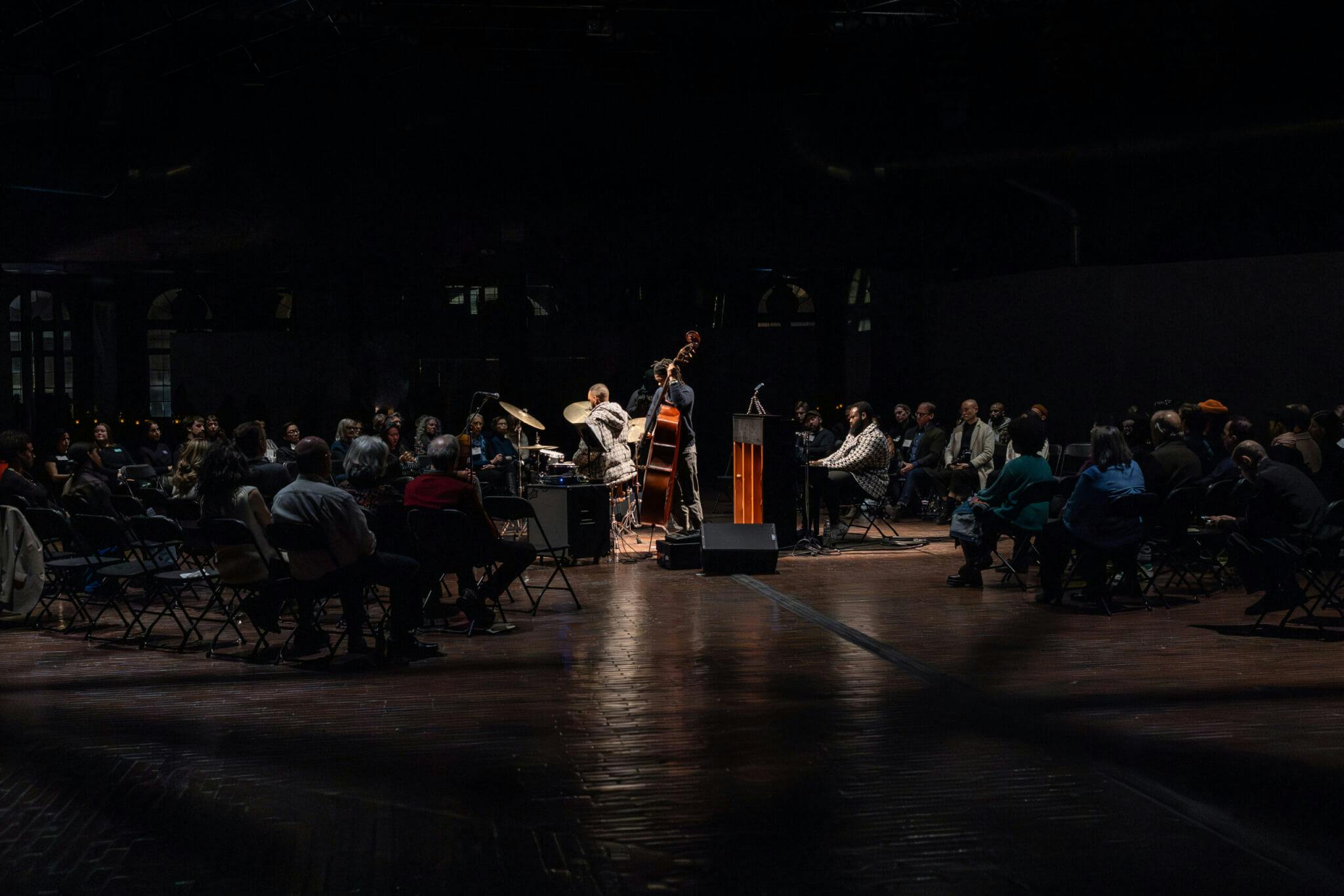 Musicians perform around a clustered audience in a darkened room.