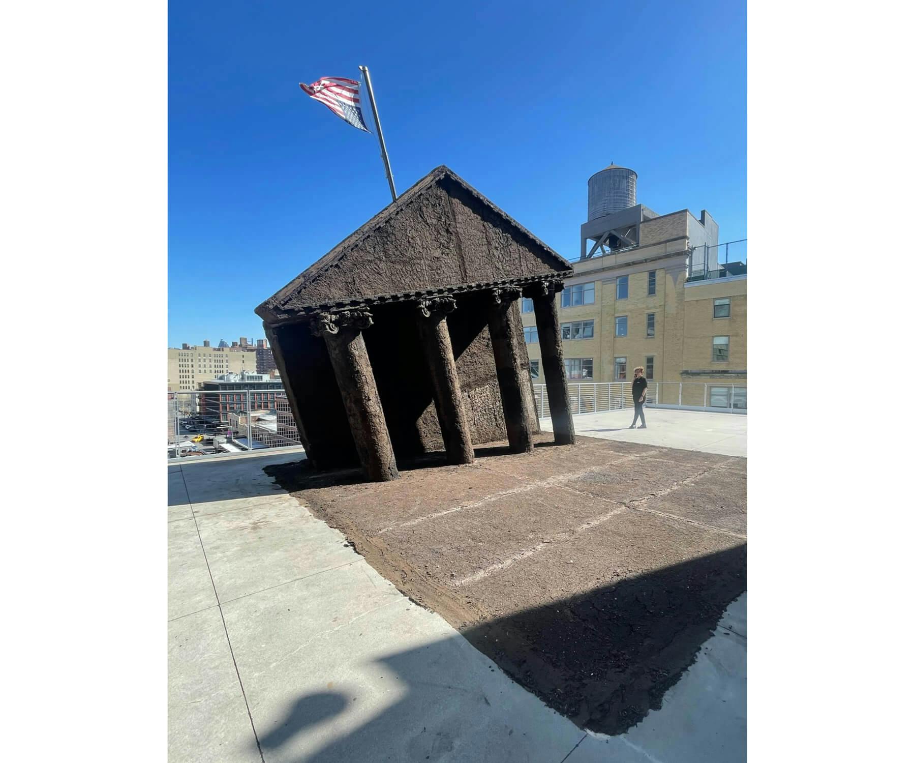 A sculpture of a slanting building with an upside down American flag pole stands in front of surrounding buildings.