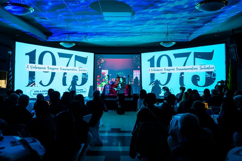 Two screens display the year "1975" while audience members sit at tables, observing the event.