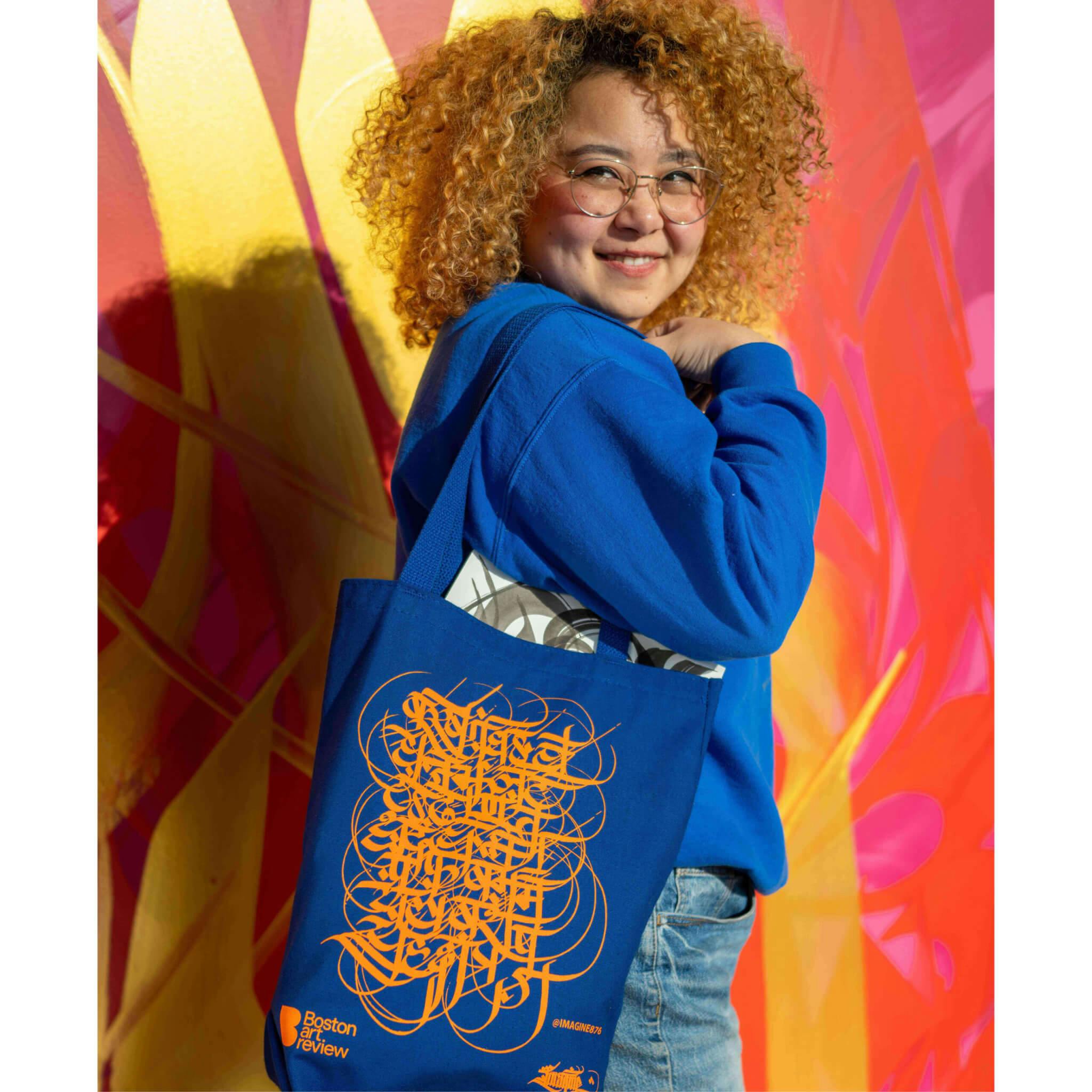 Sneha Shrestha, a young medium-light skinned woman with blonde hair, smiles while modeling the blue and orange tote bag she designed with Boston Art Review.