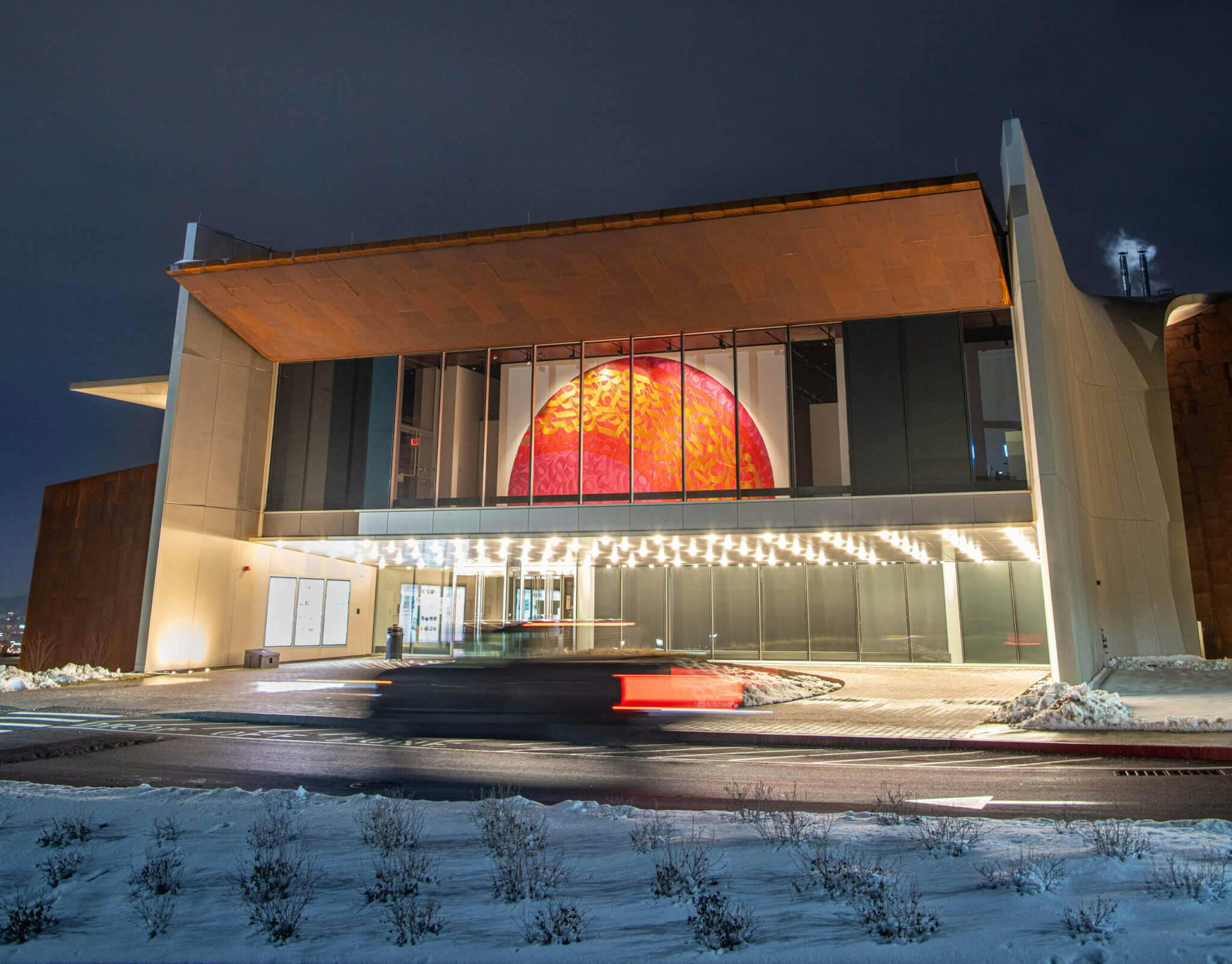 A rectangular museum exterior surrounded by small patches of snow. A ceiling of light-bulbs illuminates the entrance.