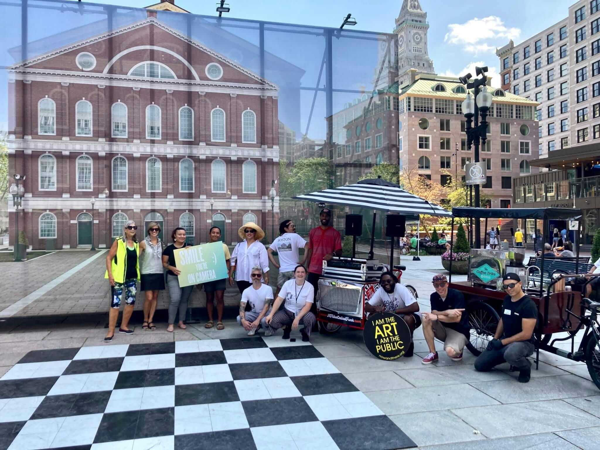 A group of visitors and staff pose in front of a scene of buildings and a black and white checker board sidewalk design.